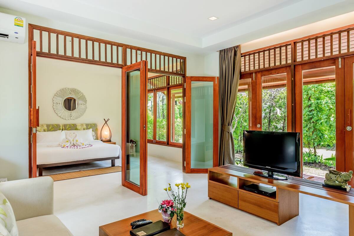 A modern hotel room with a bed, a TV, a coffee table, and a view of greenery through large windows.