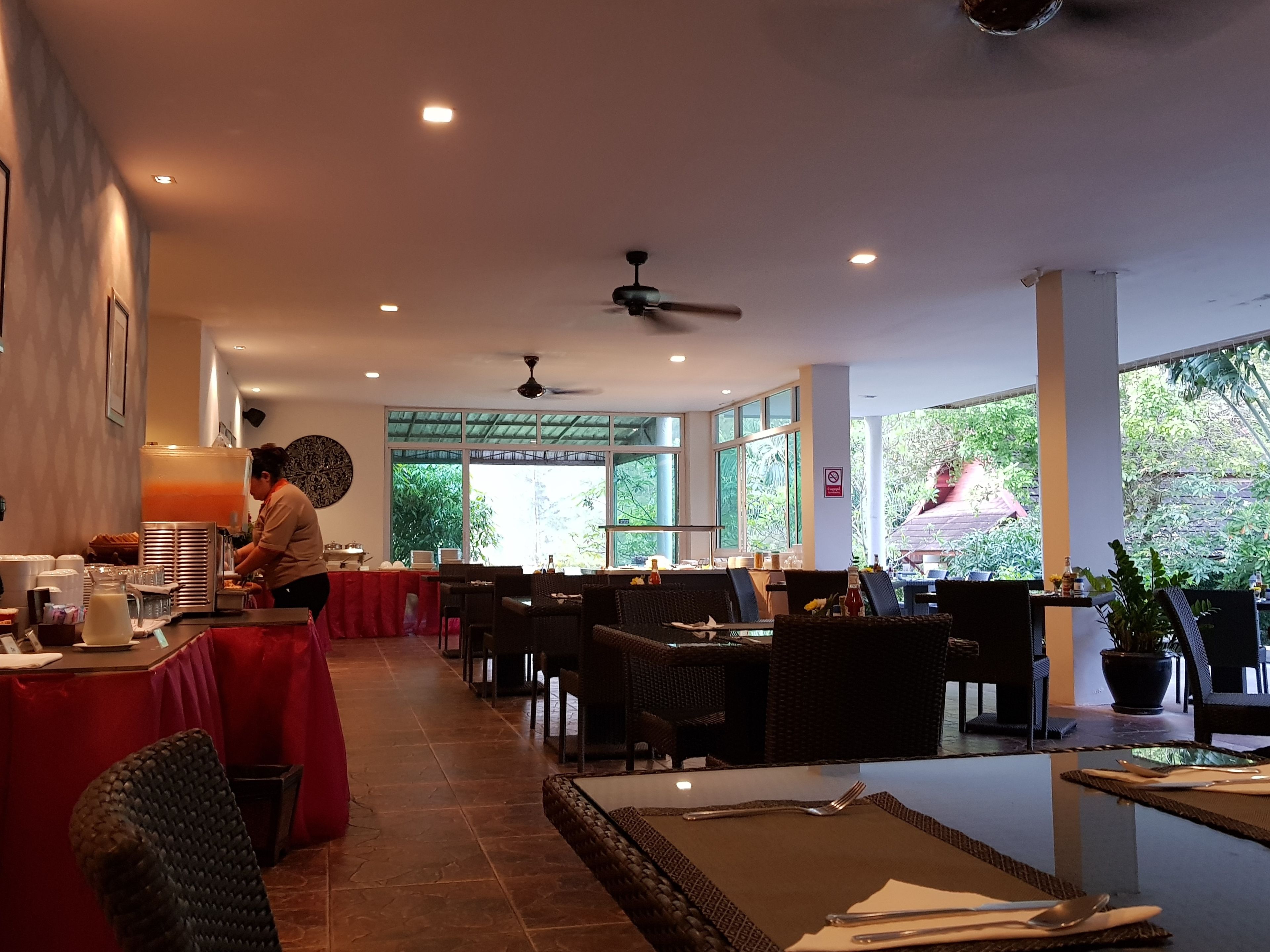 A restaurant with tables and chairs, a counter with a server, and large windows with a view of greenery.