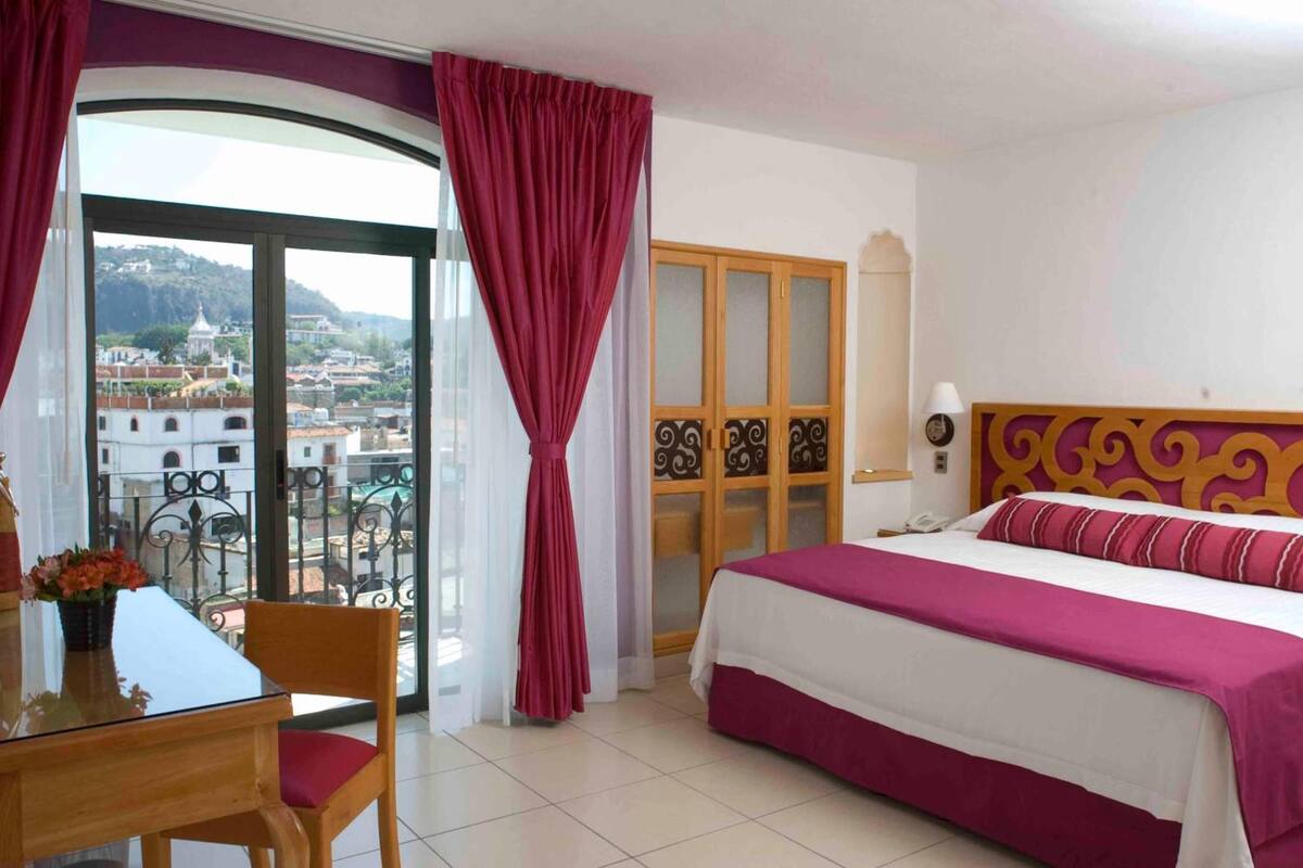 A hotel room with a bed, a desk with a chair, a balcony with a view of the city, and a door with a decorative panel.