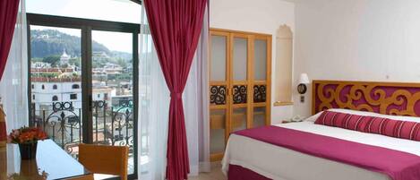 A hotel room with a bed, a desk with a chair, a balcony with a view of the city, and a door with a decorative panel.