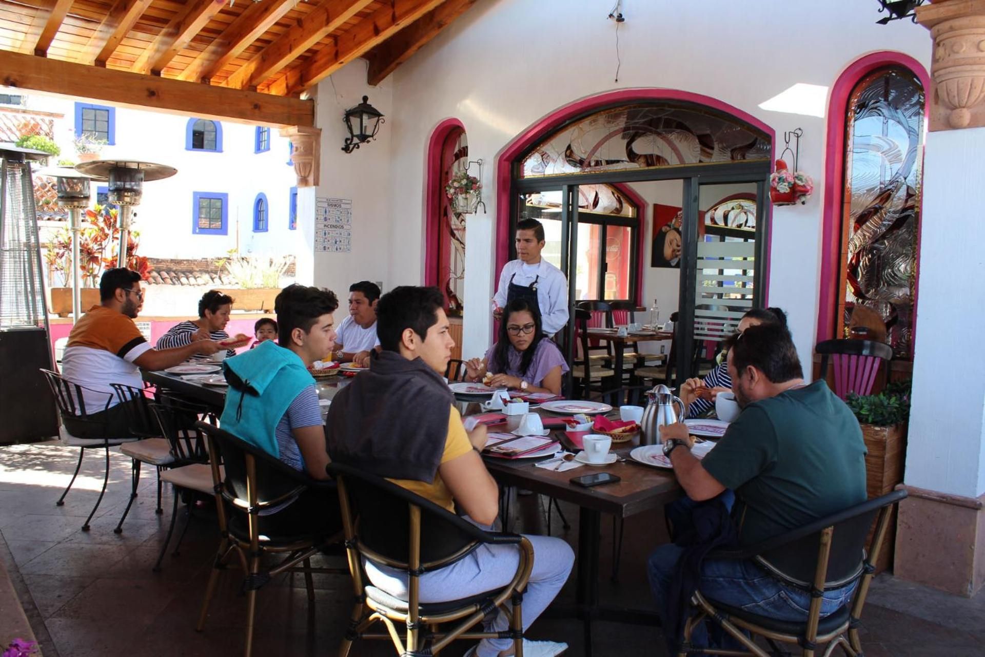 A group of people dining at an outdoor café with tables and chairs, a waiter attending to them, and a building with arched windows in the background.
