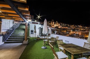 Una terraza en la azotea con una escalera de madera, asientos exteriores y vistas a un paisaje urbano nocturno.