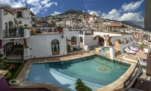 A pool area with a sun-shaped design, surrounded by white buildings with balconies and a view of a hillside town.