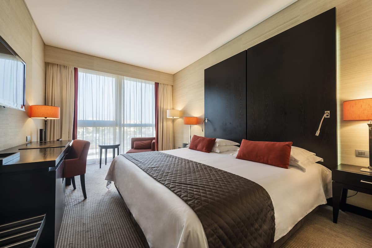 A hotel room with a large bed, a desk with a chair, a small table, and two lamps.
