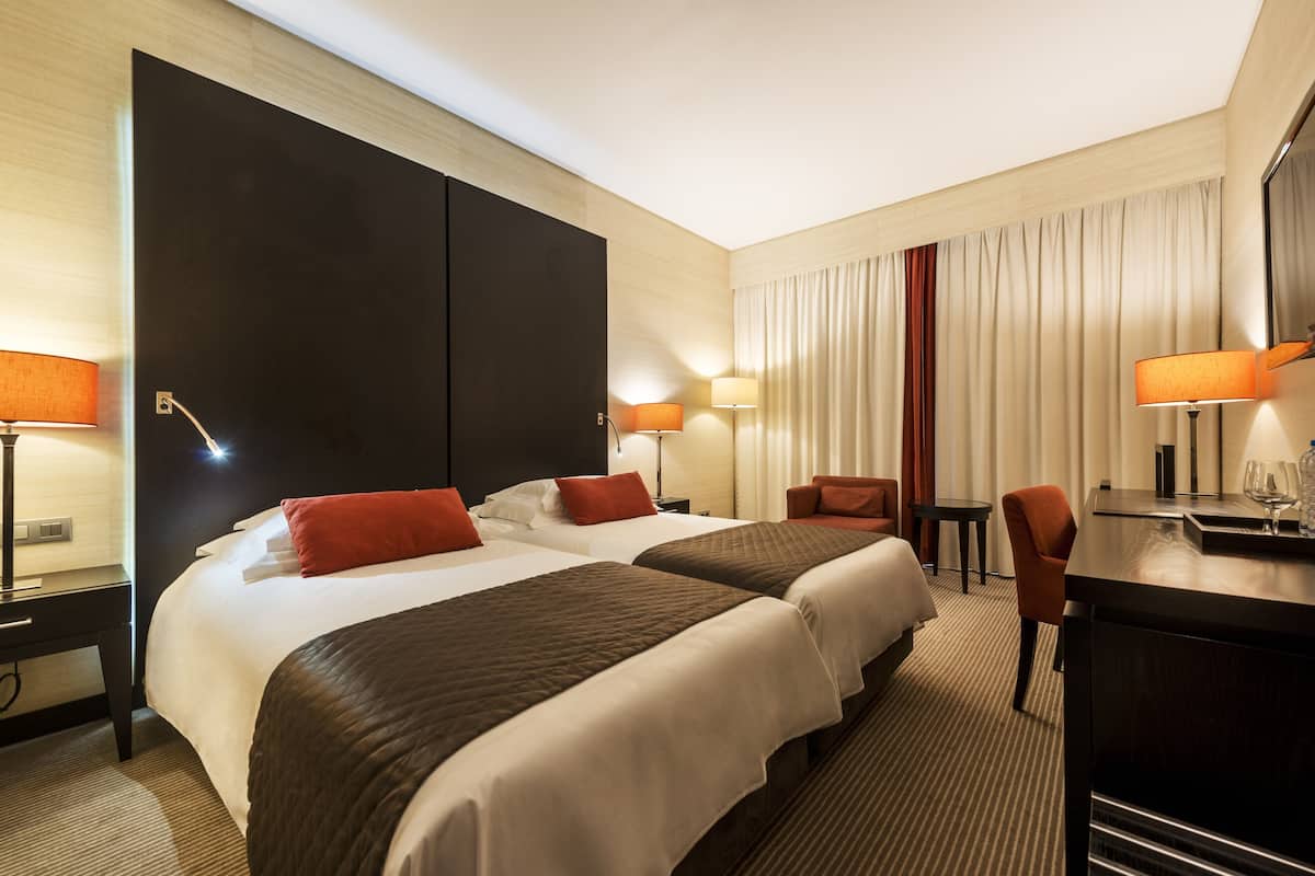 A hotel room with two beds, a desk, a chair, and a large window with curtains.