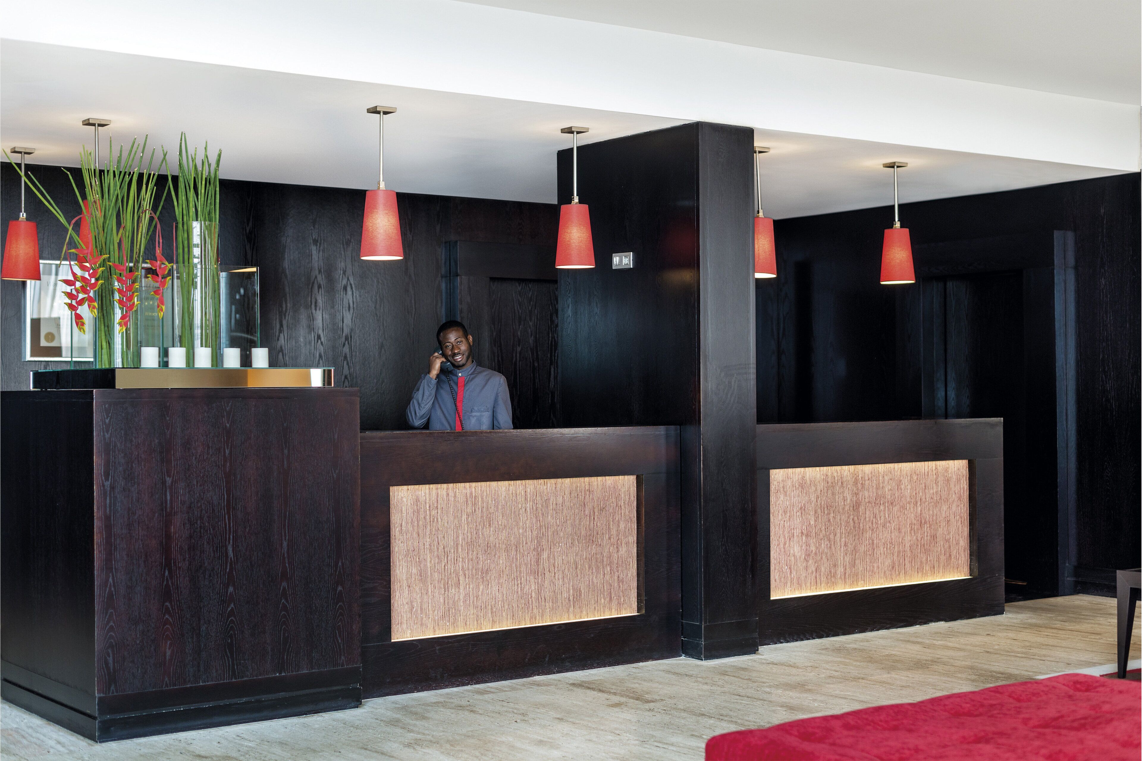 A reception area with a dark wood reception desk, red pendant lights, and a vase with red flowers.