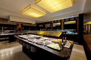 A buffet area with various food items displayed on a counter, overhead lighting, and a view of the city at night. - Hotel Baía (Luanda)