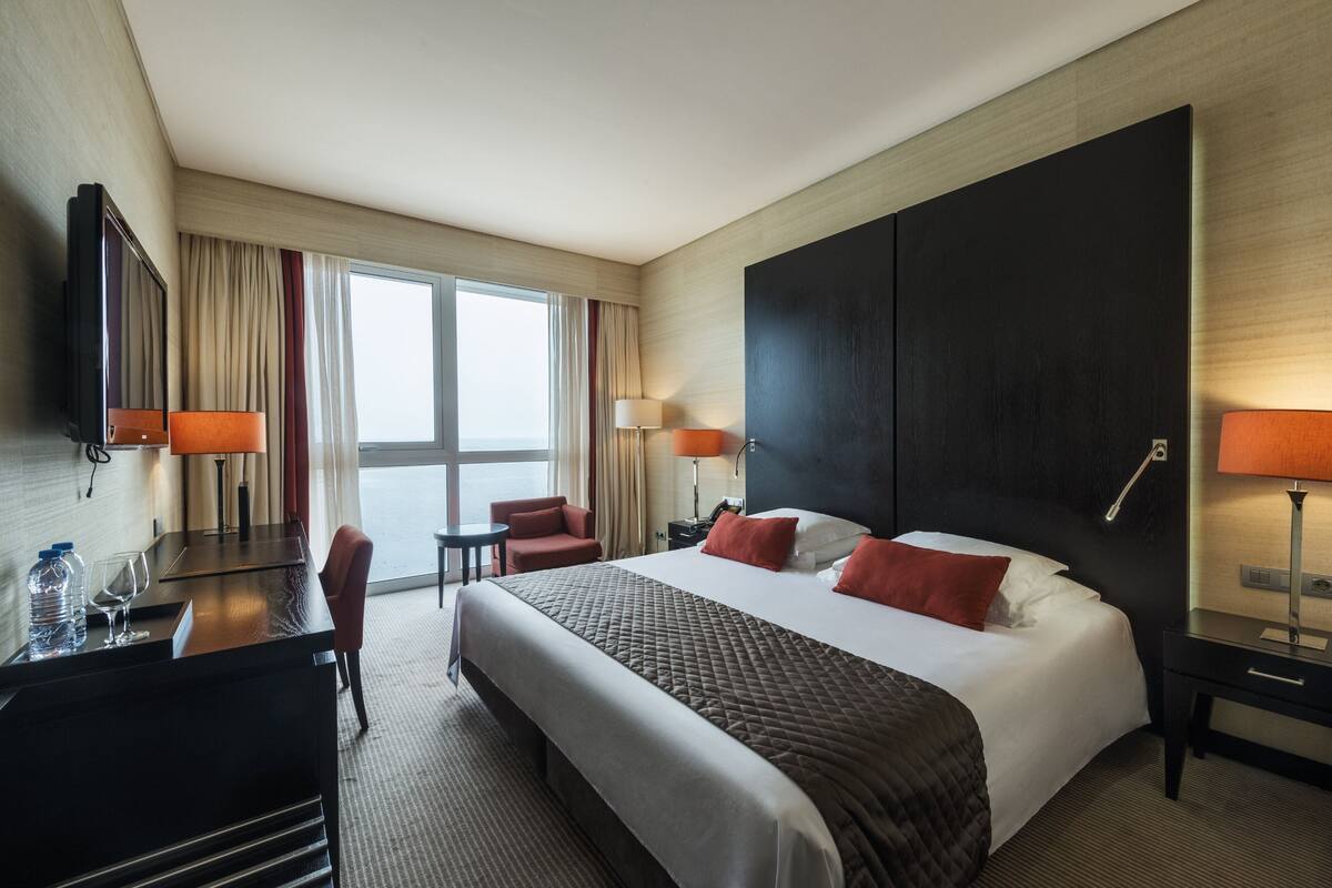 A hotel room with a large bed, two bedside tables, a desk with a chair, a TV, and a window with curtains.