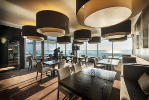Un comedor moderno con varias mesas y sillas, lámparas colgantes grandes y vistas del paisaje urbano a través de amplios ventanales.