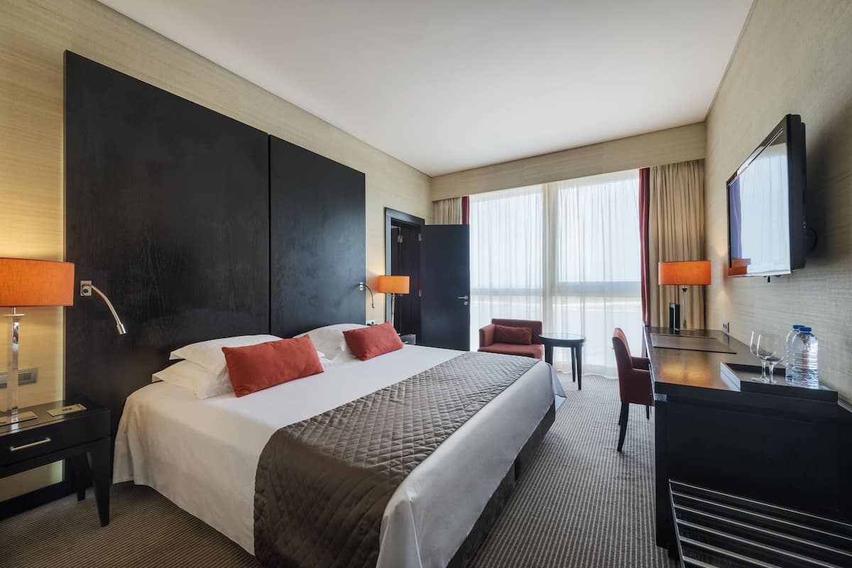 A hotel room with a large bed, two bedside tables, a desk with a chair, a TV, and a window with curtains.
