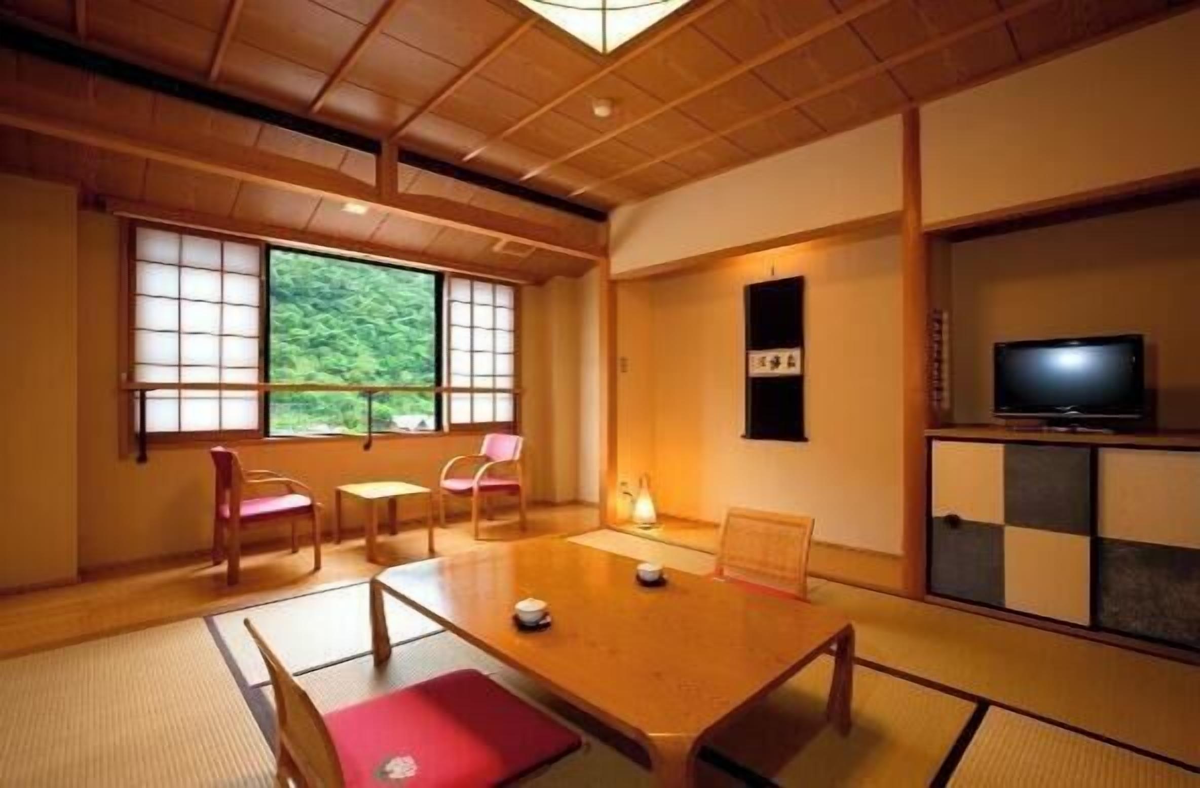 A traditional Japanese room with tatami flooring, a low table, chairs, a television, and a view of greenery outside.