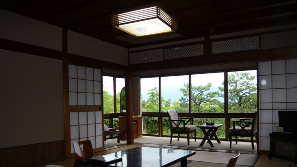 Una habitación japonesa tradicional con suelo de tatami, una mesa baja, sillas y vistas a la vegetación a través de amplias ventanas.