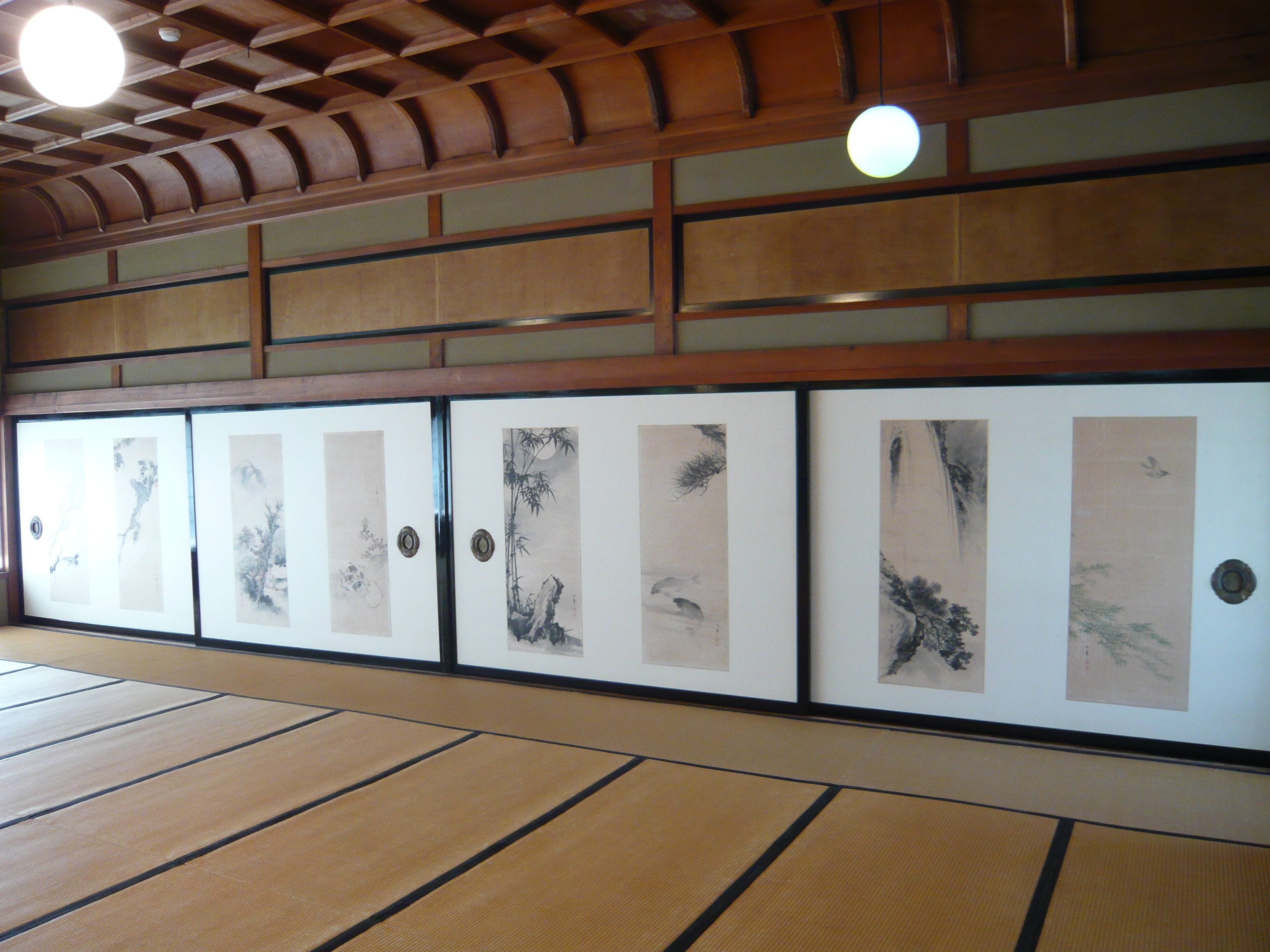 Una habitación japonesa tradicional con suelo de tatami, puertas correderas decoradas con obras de arte y techo de madera.