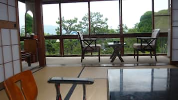 A room with tatami flooring, a low table, and two chairs. Large windows offer a view of trees and hills.