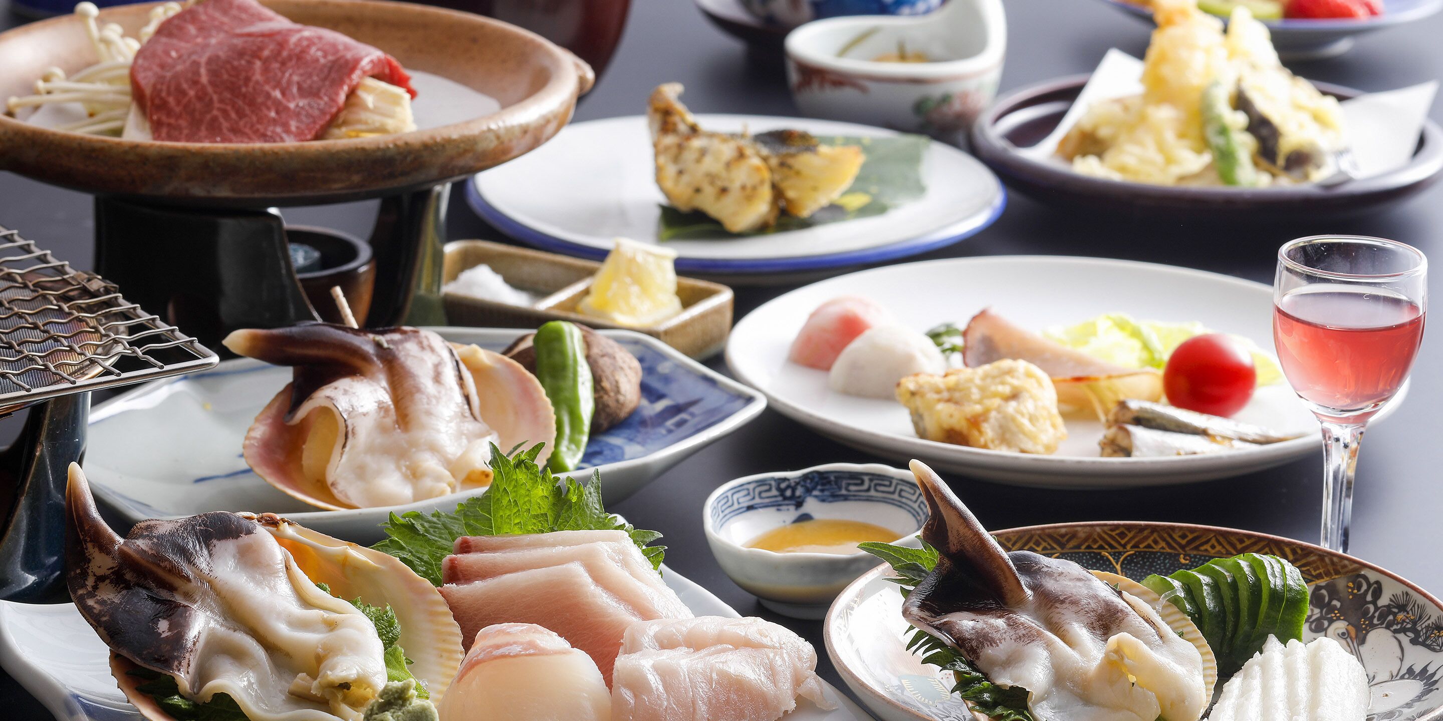 A variety of seafood dishes on plates, a glass of pink drink, and a traditional Japanese hot pot setup.