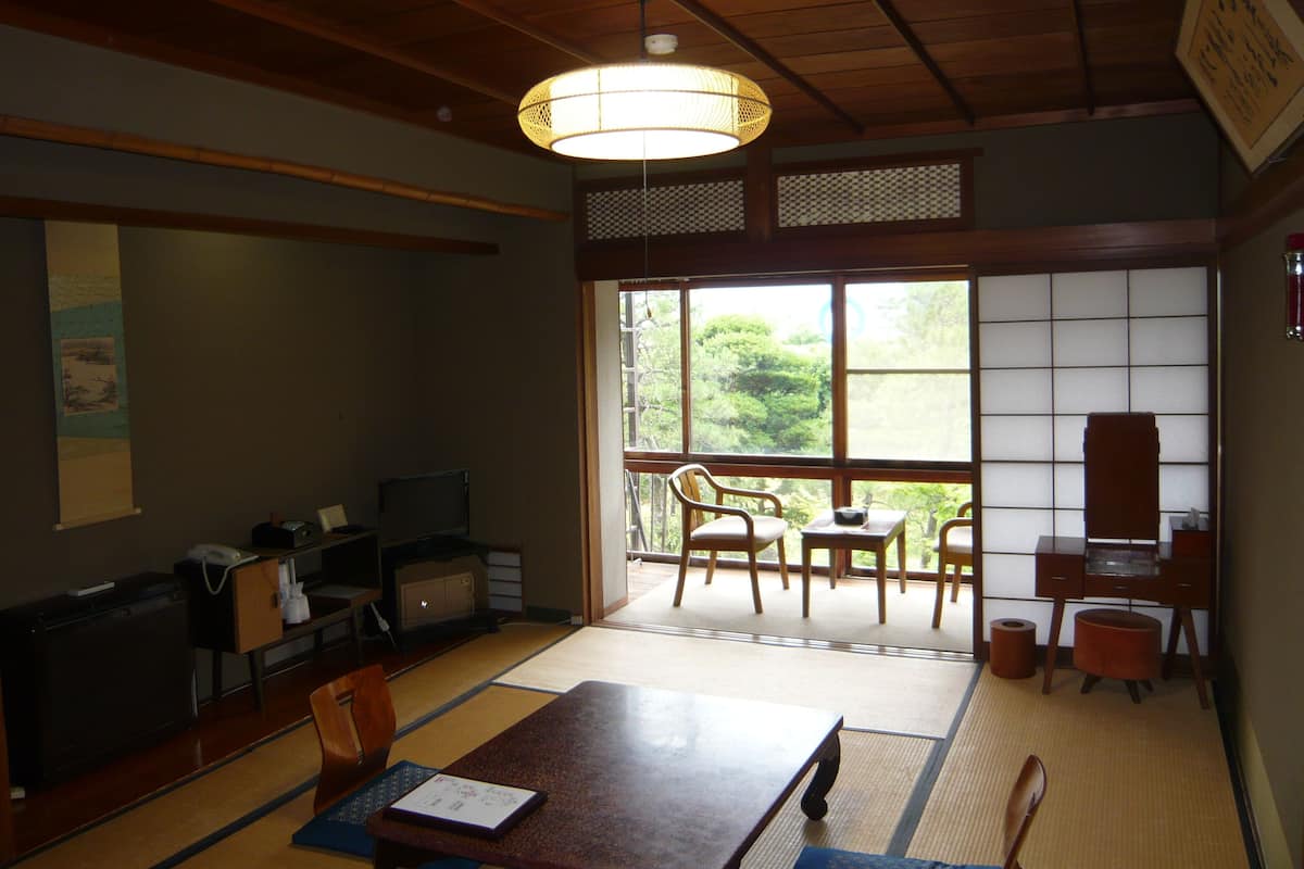 Una habitación japonesa tradicional con suelo de tatami, una mesa baja, sillas, un escritorio con una computadora y un ventanal con vistas a zonas verdes.
