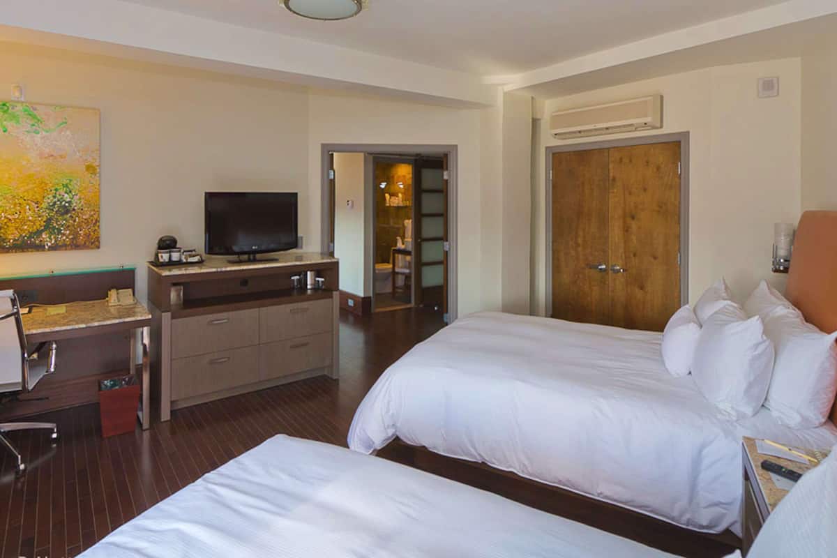 A hotel room with two beds, a TV on a wooden dresser, a painting on the wall, and a bathroom visible through an open door.