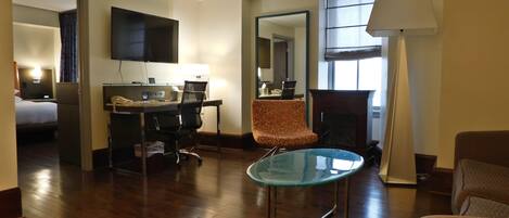 A hotel room with a wooden floor, a glass table, a desk with a computer, a chair, a bed, a TV, and a fireplace.