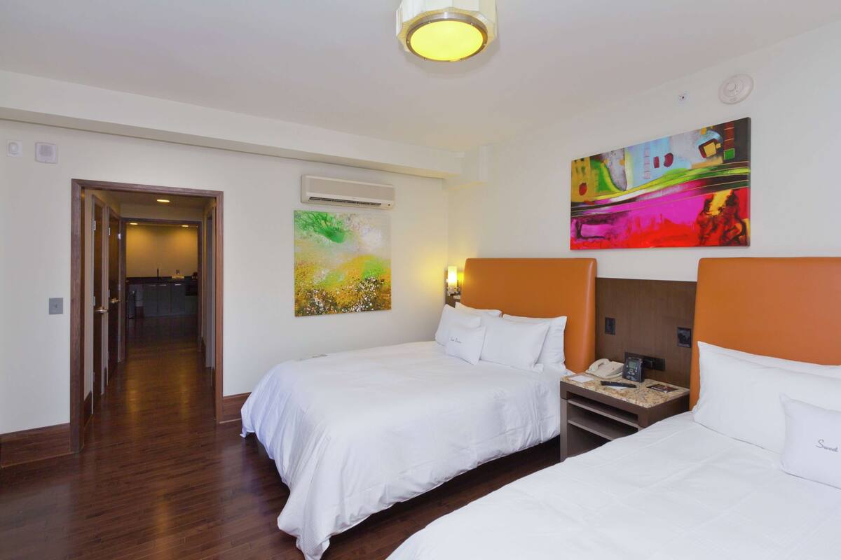 A hotel room with two beds, a painting on the wall, and a door leading to another room.