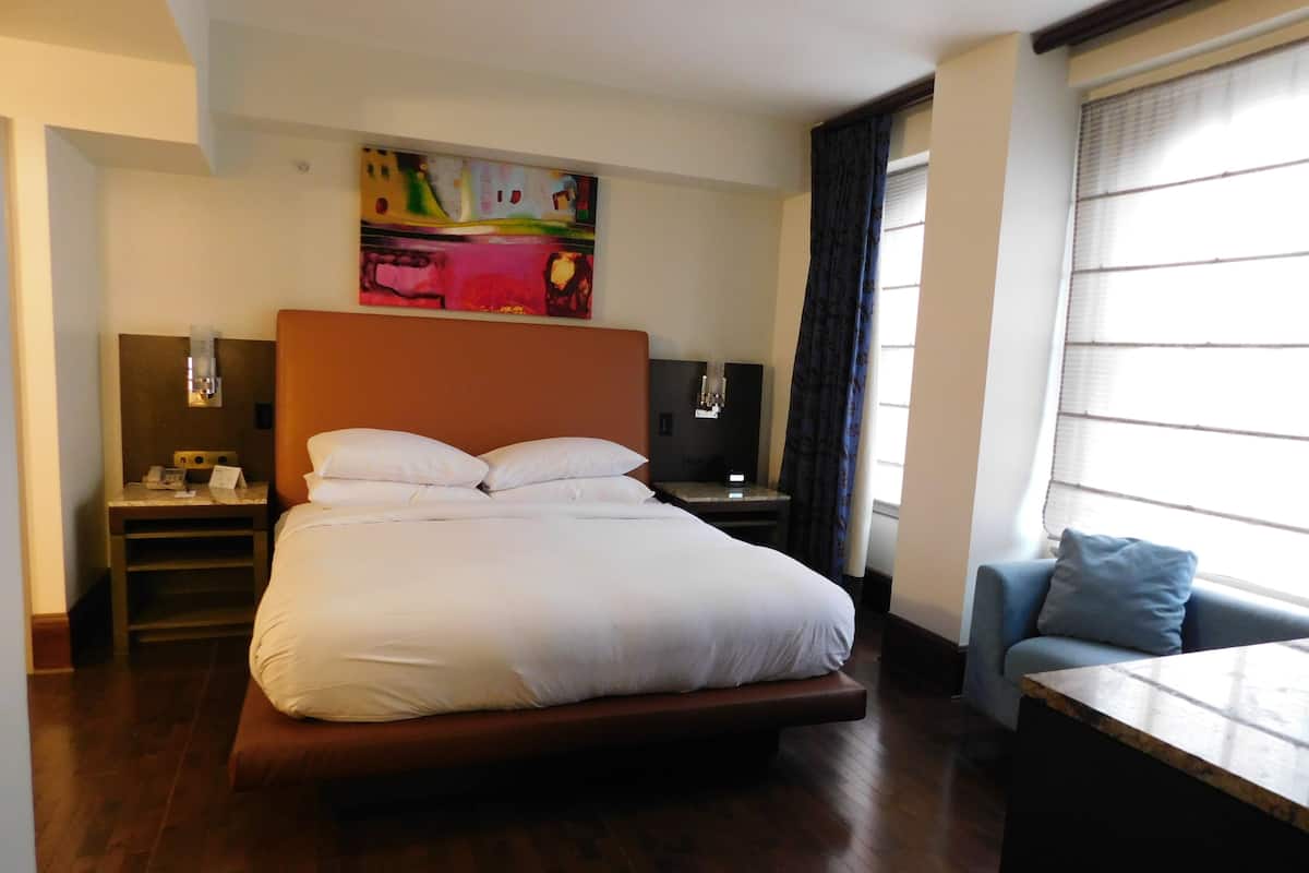 A hotel room with a large bed, a desk, a chair, a nightstand, a painting on the wall, and a window with curtains.