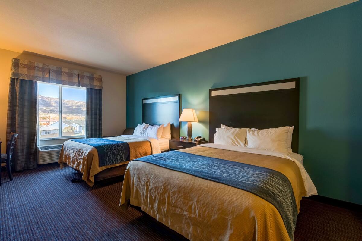 A hotel room with two beds, a large window with curtains, a view of a mountainous landscape, and a nightstand with a lamp.
