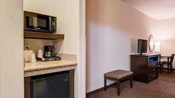 Habitación de hotel compacta con kitchenette, que incluye microondas, cafetera y refrigerador. Cuenta con una pequeña zona de estar con una silla y un banco, y un televisor sobre un mueble.