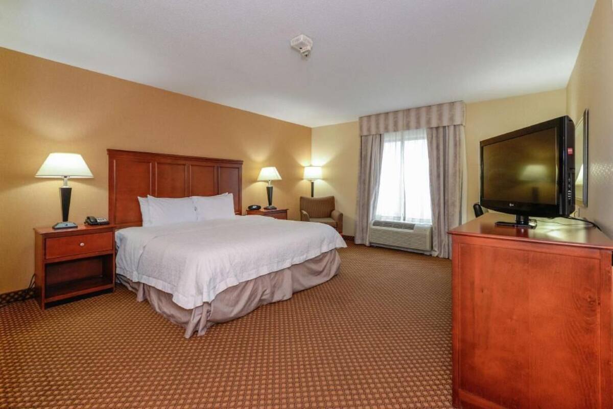 Ett hotellrum med en säng, två sänglampor, en tv, ett skrivbord och ett fönster med gardiner.