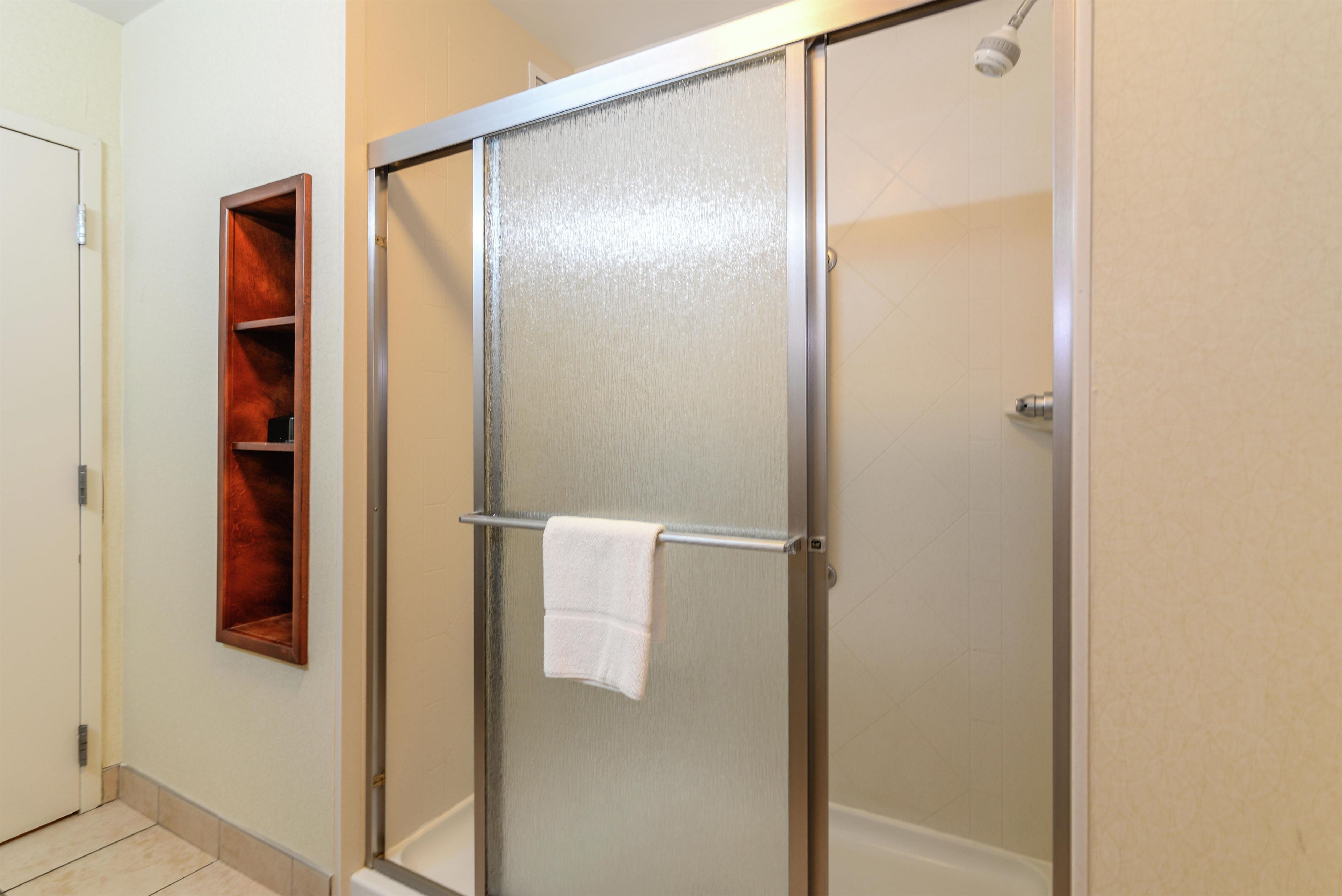 A shower with a glass door, a towel hanging on the door handle, and a wooden shelf to the left.