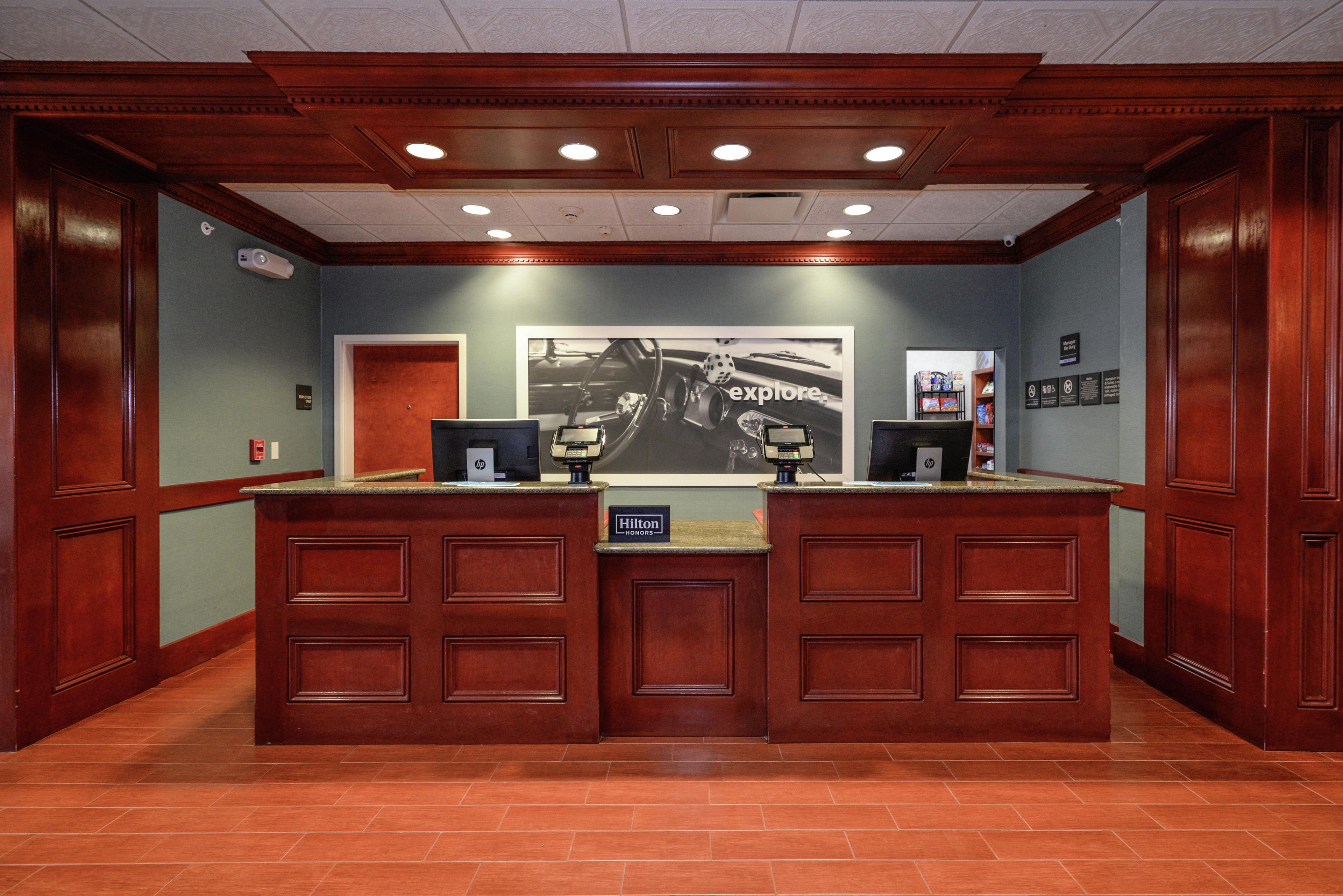 A hotel reception desk with a large framed advertisement, a digital screen, and a sign indicating 'Manager on Duty'.