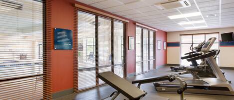 Un gimnasio con equipo de ejercicio, incluyendo cintas de correr y una banca, y ventanas amplias con persianas.