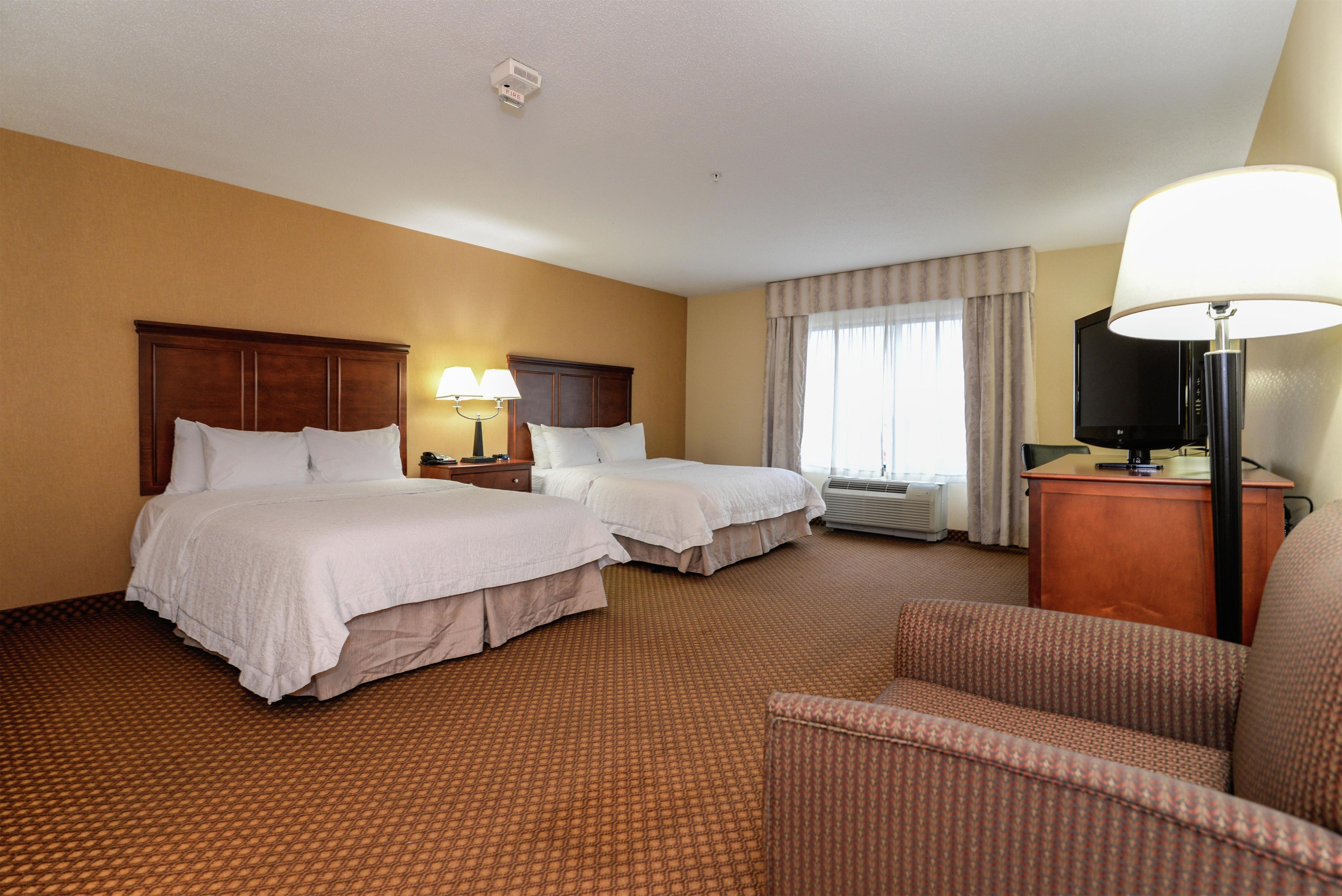A hotel room with two beds, a desk with a lamp, a television, and an air conditioning unit.