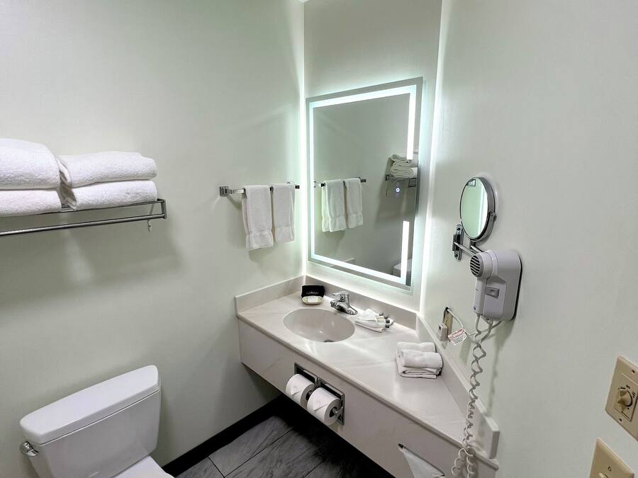 Un baño moderno con inodoro, lavabo y espejo. Hay toallas en un estante y un secador de pelo en la pared.