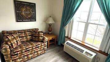 A living room with a patterned sofa, a table with a lamp, a wall-mounted decorative piece, and a window with curtains.