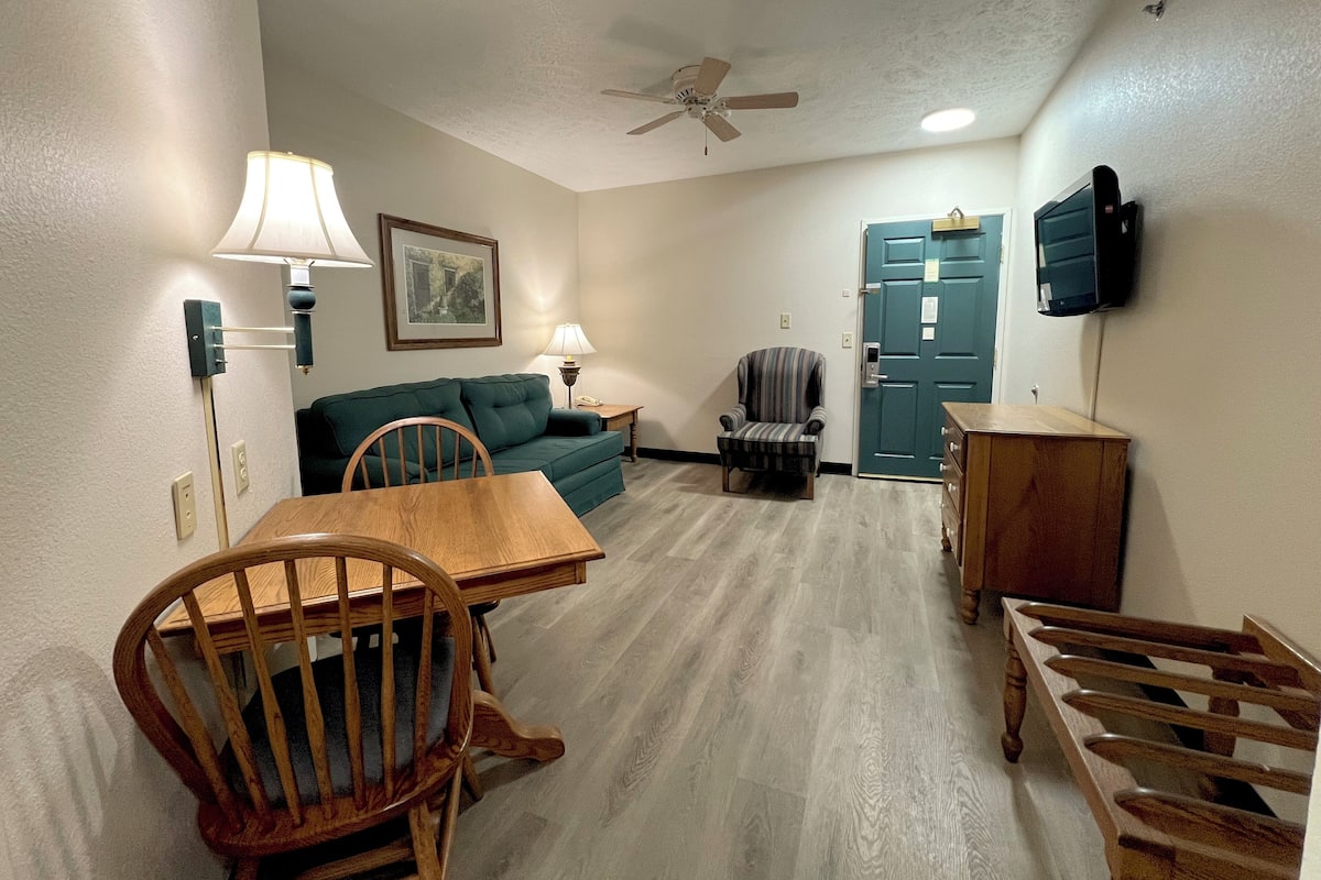 A compact living space with a dining table, a sofa, a chair, a TV mounted on the wall, and a ceiling fan.