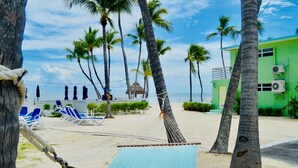A beach with a hammock, palm trees, lounge chairs, and a green building with air conditioning units.