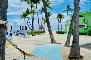 A beach with a hammock, palm trees, lounge chairs, and a green building with air conditioning units.