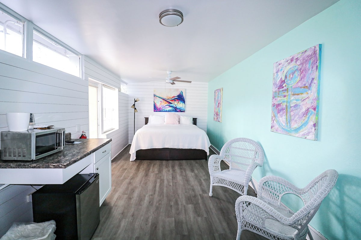 A bedroom with a bed, a desk with a microwave, a chair, and artwork on the walls.