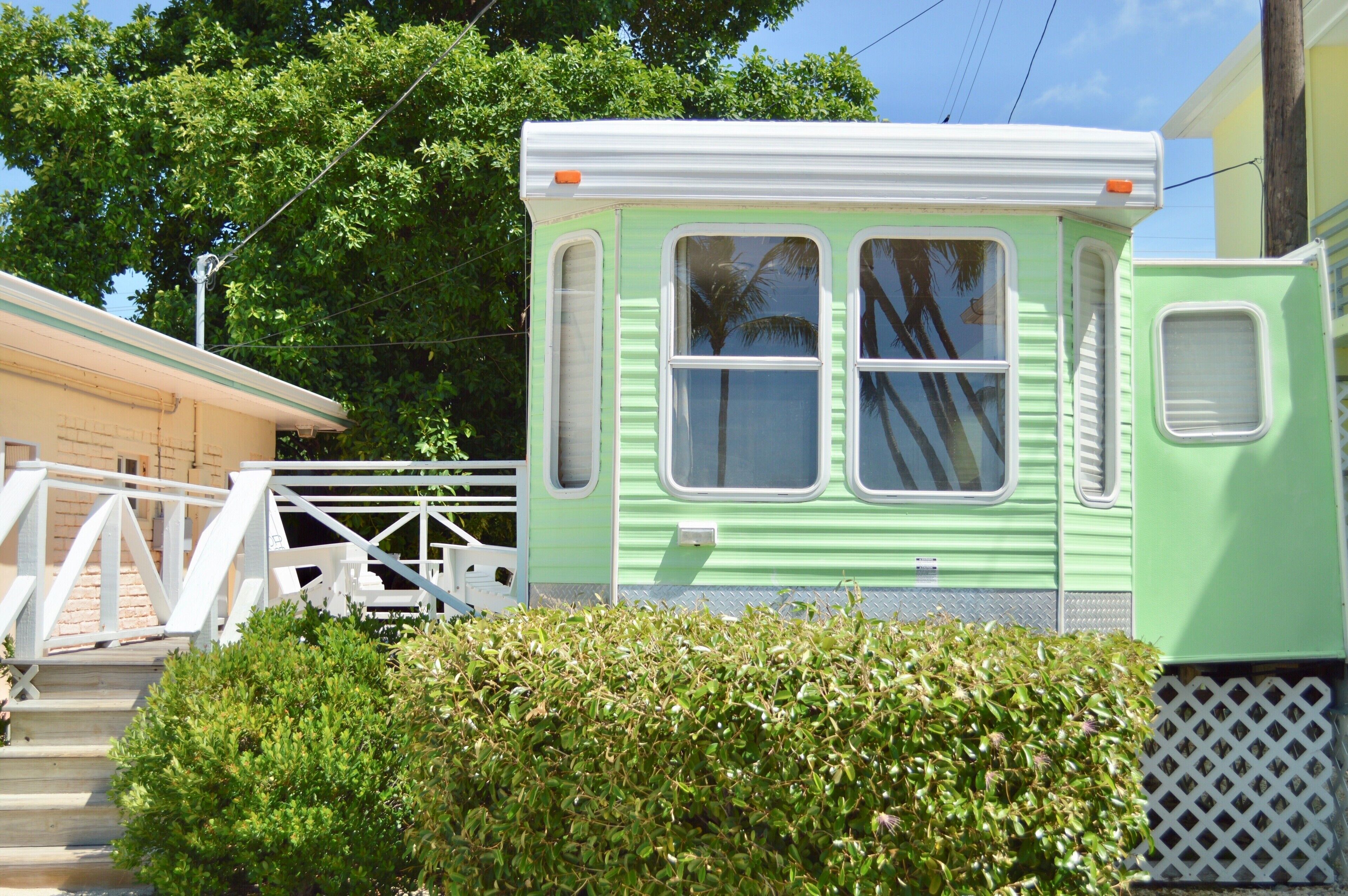 Une maison mobile verte, avec une porte et des fenêtres blanches, un petit porche et une haie au premier plan.