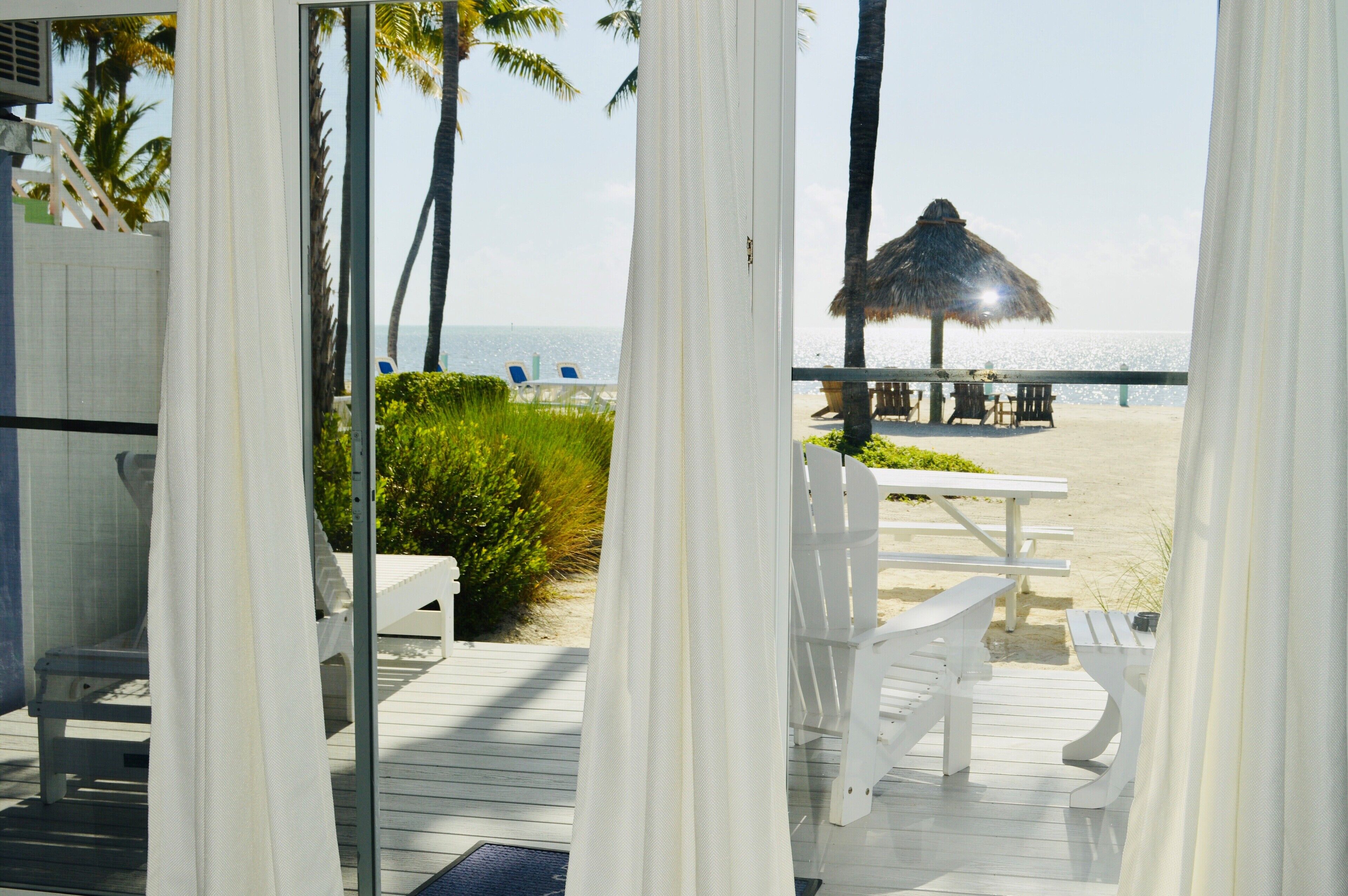 A beach view from a balcony with white curtains, a thatched umbrella, and white outdoor furniture.