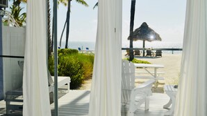 A beach view from a balcony with white curtains, a thatched umbrella, and white outdoor furniture.