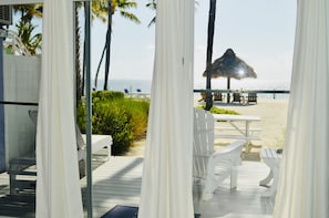 A beach view from a balcony with white curtains, a thatched umbrella, and white outdoor furniture.