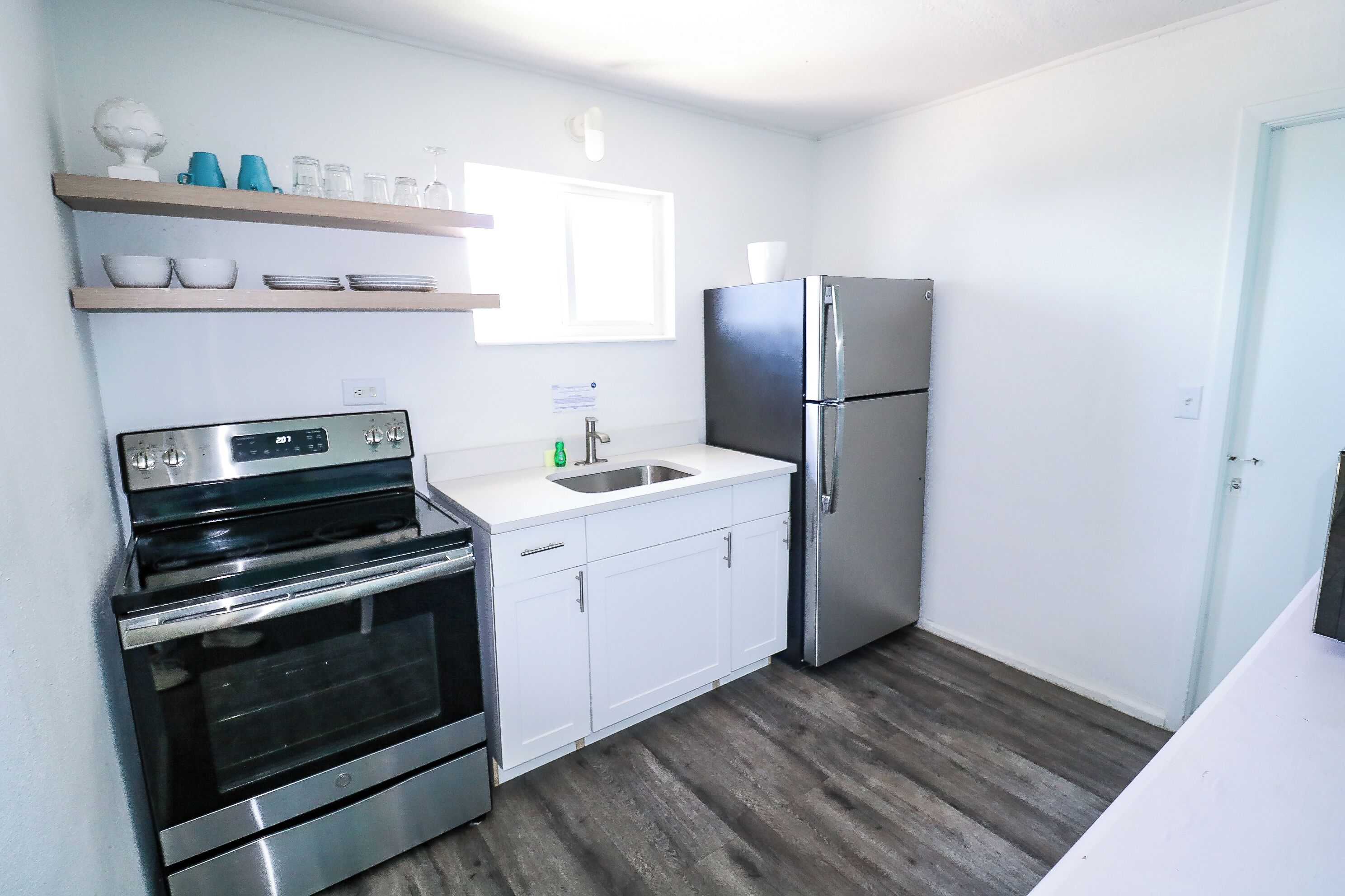 Cocina compacta con armarios blancos, horno de acero inoxidable, refrigerador y piso de madera.