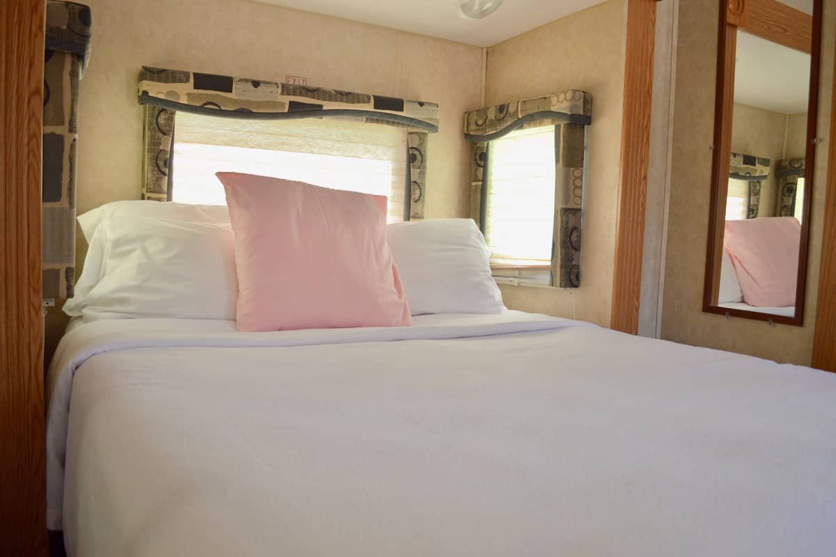 A neatly made bed with white and pink linens, a wooden headboard, and a mirror on the wall.