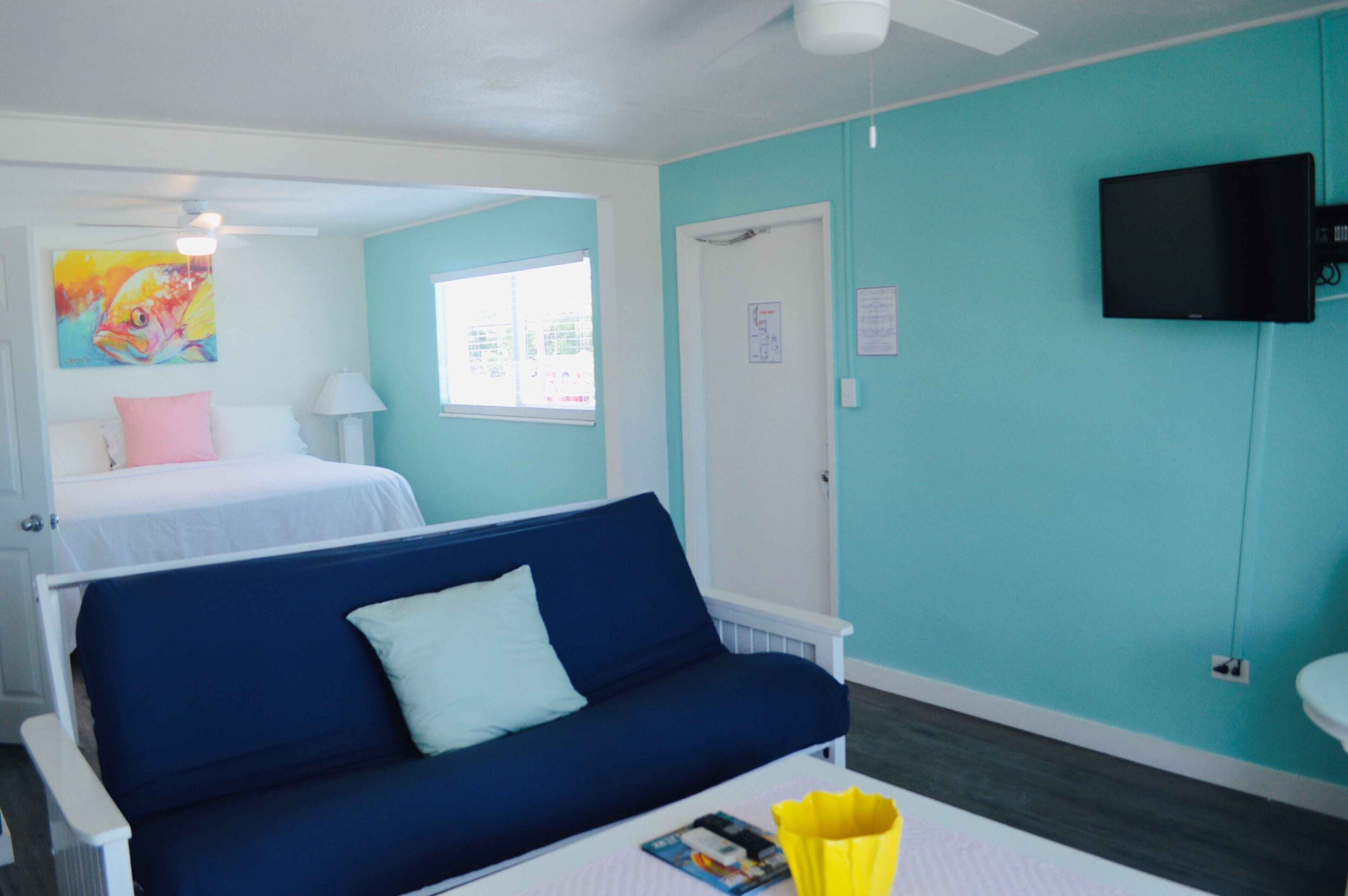A room with a bed, a blue sofa, a small table with a magazine, a TV mounted on the wall, and a window with blinds.