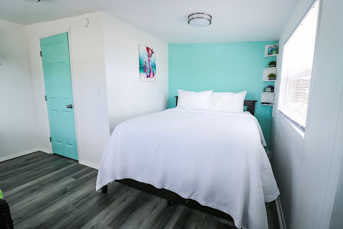 A bedroom with a bed, a teal door, a white wall, and a window.