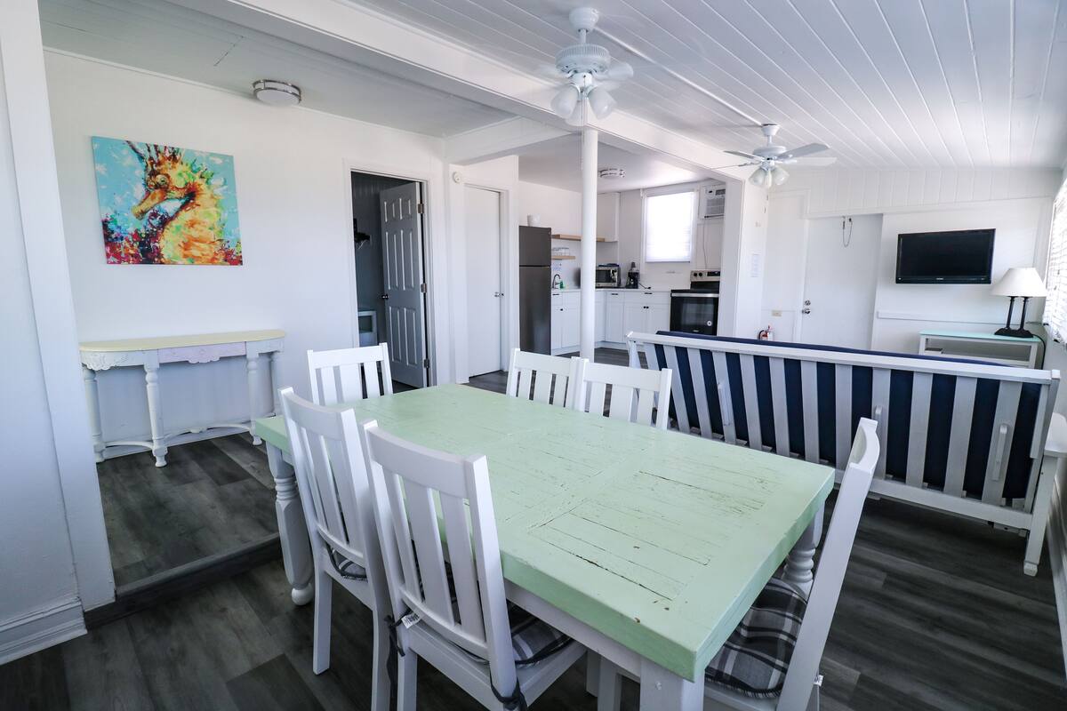 A dining area with a green table and white chairs, a ceiling fan, a kitchen in the background, and a colorful painting on the wall.