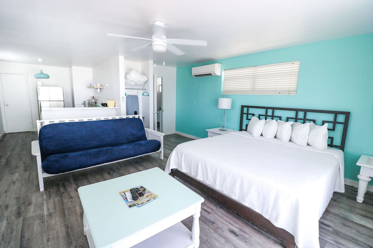 A hotel room with a bed, a blue sofa, a small table, a ceiling fan, a white nightstand, and a bathroom visible through an open door.
