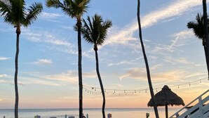 A beach with white lounge chairs, a thatched umbrella, and palm trees under a clear sky.