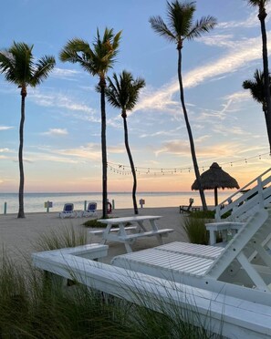A beach with white lounge chairs, a thatched umbrella, and palm trees under a clear sky.