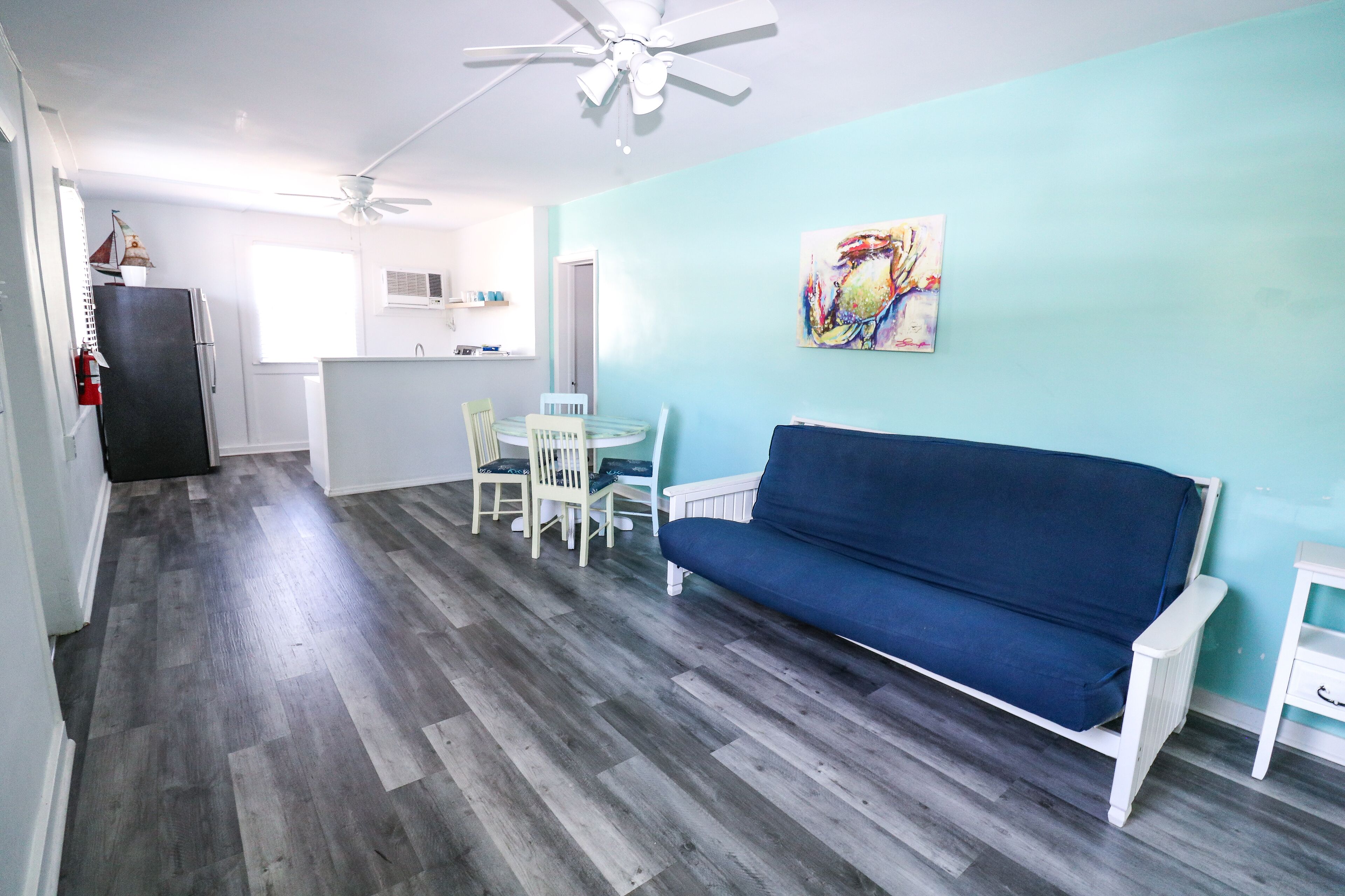 A compact living space with a blue sofa, a dining table with chairs, a kitchen area, and a ceiling fan.